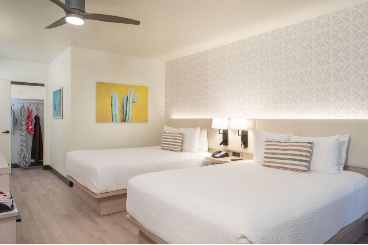 A hotel room with two beds, a ceiling fan, a wall-mounted phone, and a painting.
