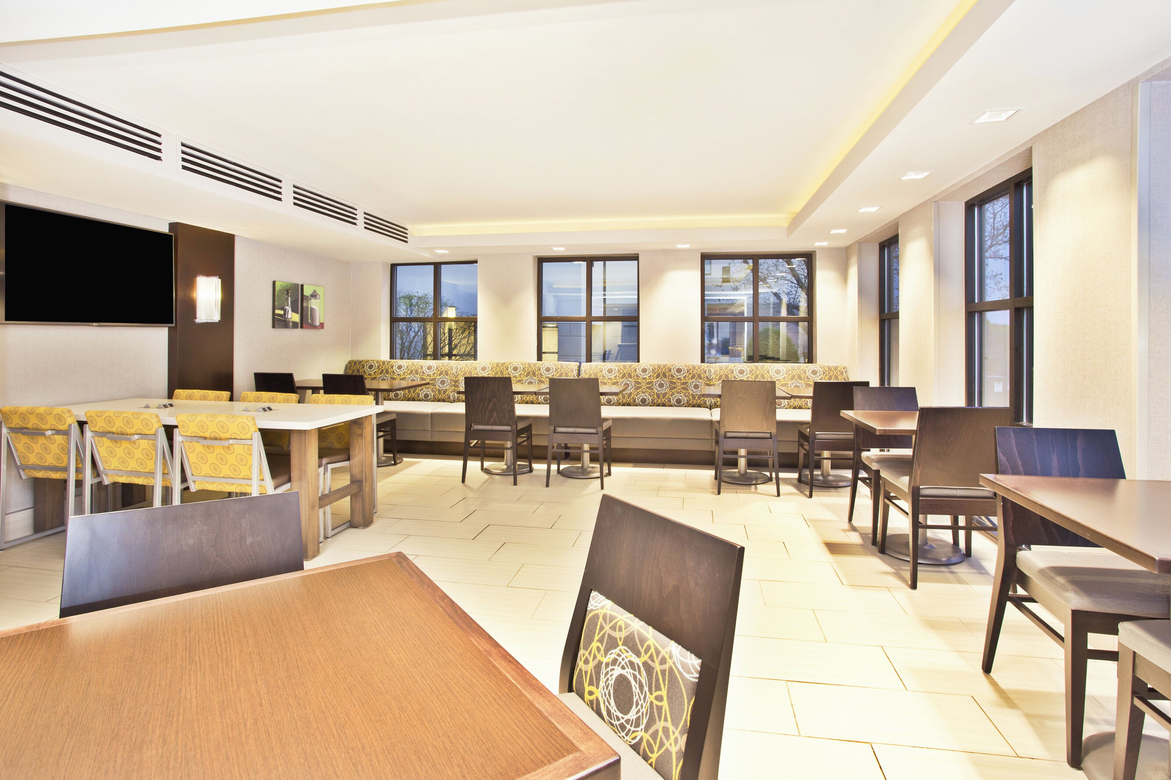 A modern dining area with multiple tables and chairs, large windows, and a flat-screen TV mounted on the wall.
