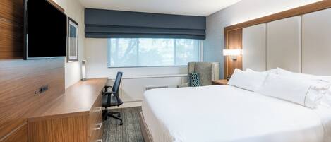 A hotel room with a large bed, a desk with a chair, a flat-screen TV, a window with a view, and a nightstand with a lamp.
