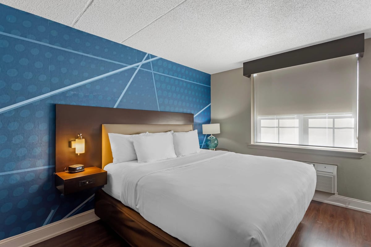 A hotel room with a large bed, a nightstand with a lamp, a wall clock, and a window with blinds.