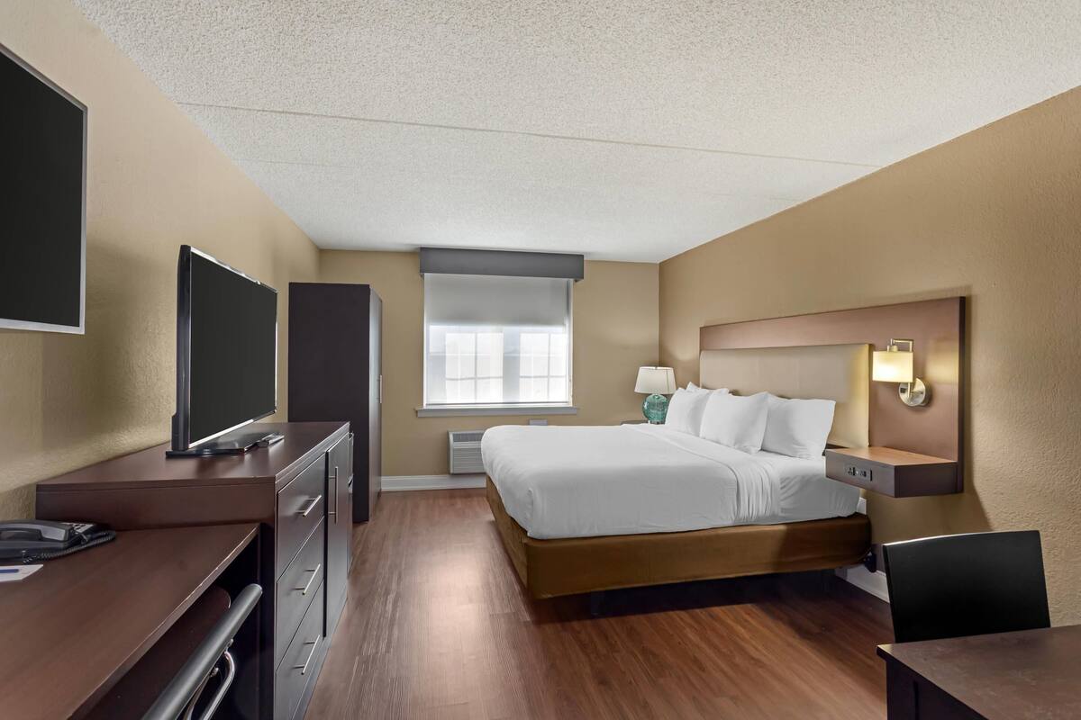 A hotel room with a bed, desk, TV, and a window with blinds.