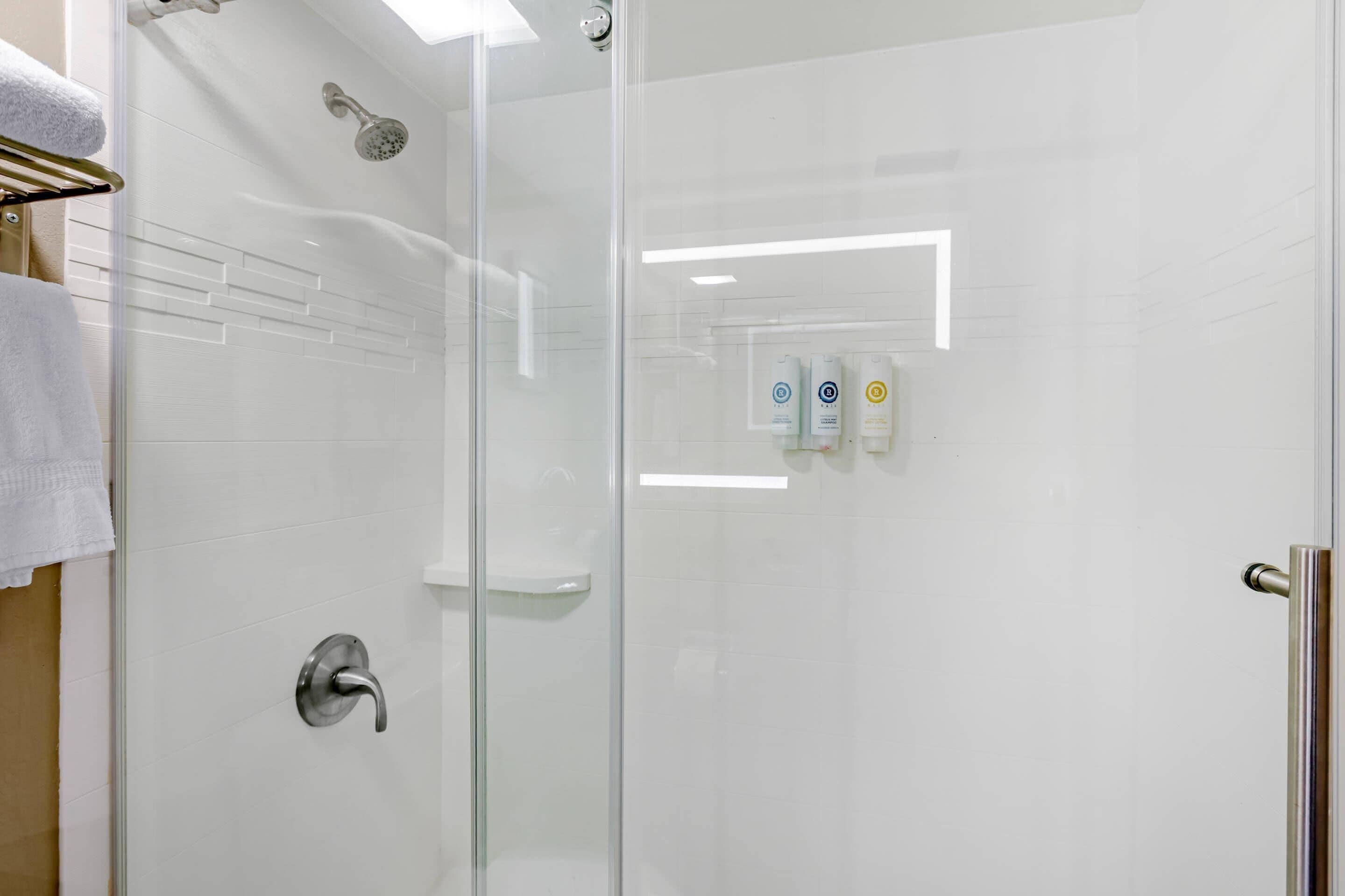 Cabina de ducha con paredes de vidrio, cabezal de ducha de mano, puerta de vidrio y un perchero con una toalla blanca.