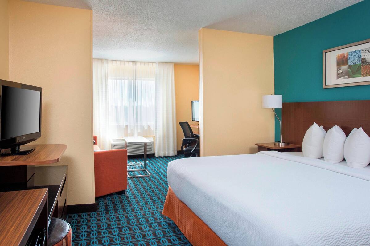 A hotel room with a bed, a desk with a television, a chair, a desk lamp, and a window with curtains.