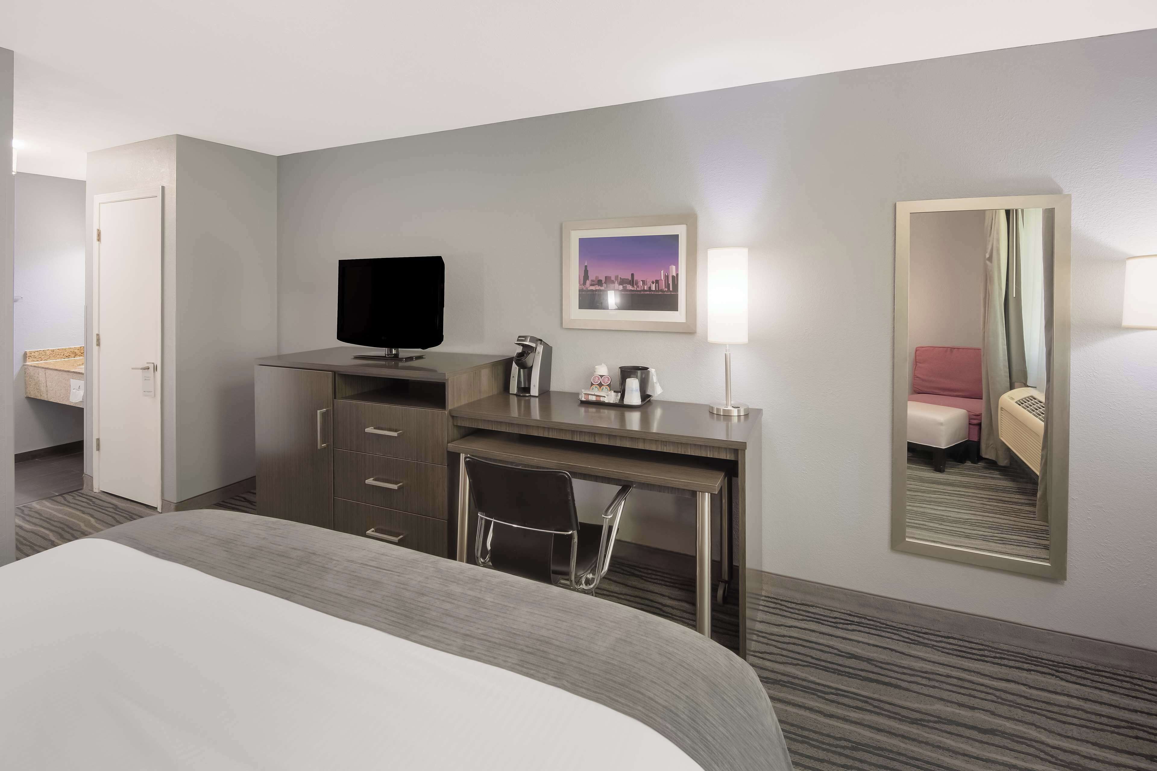 A hotel room with a bed, a desk with a chair, a television, a framed picture, a mirror, and a door.