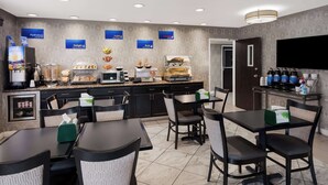 A hotel dining area with multiple tables and chairs, a self-service breakfast bar, and a television mounted on the wall.
