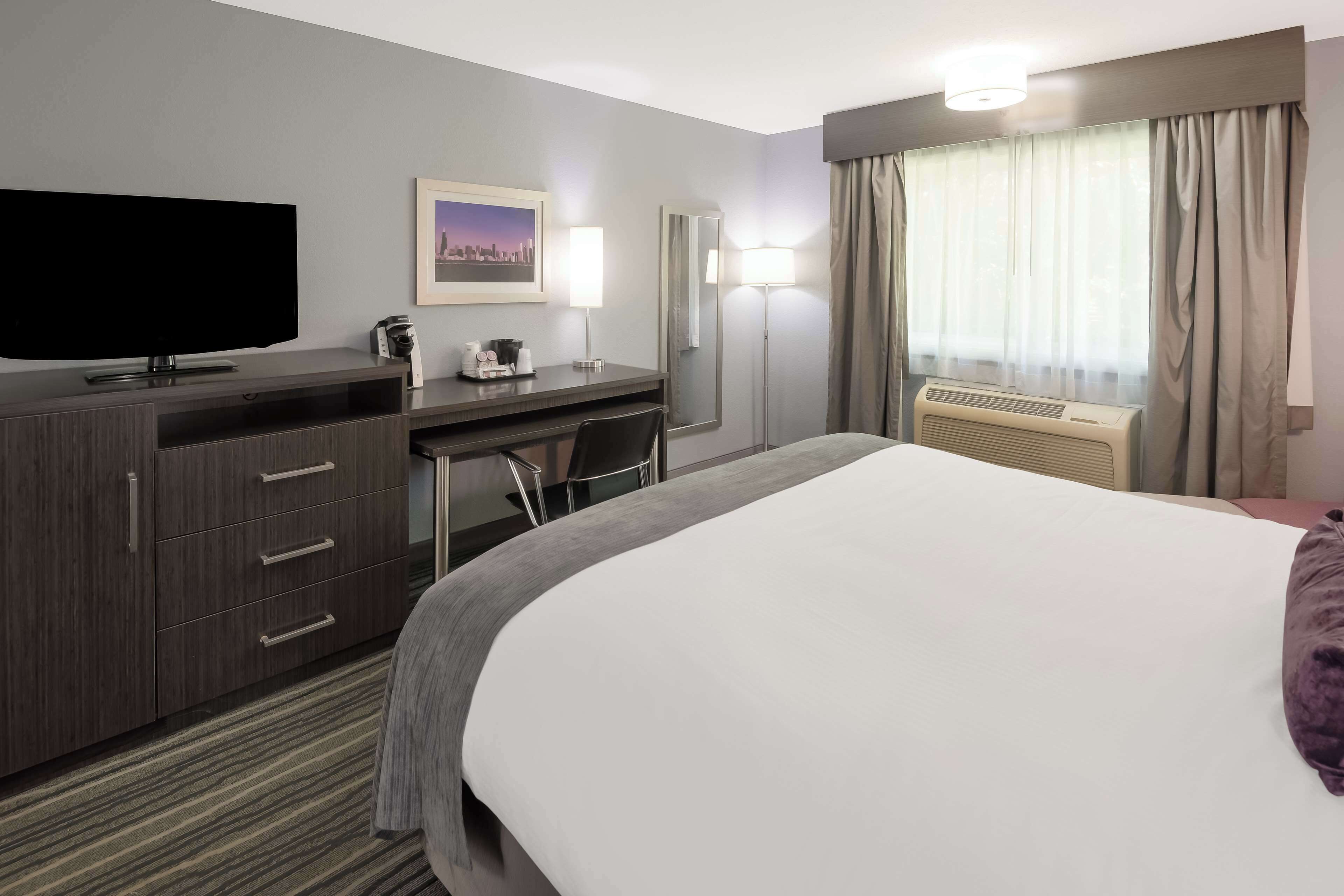 Habitación de hotel con cama, escritorio, televisor de pantalla plana, cafetera, lámpara, espejo y aire acondicionado.