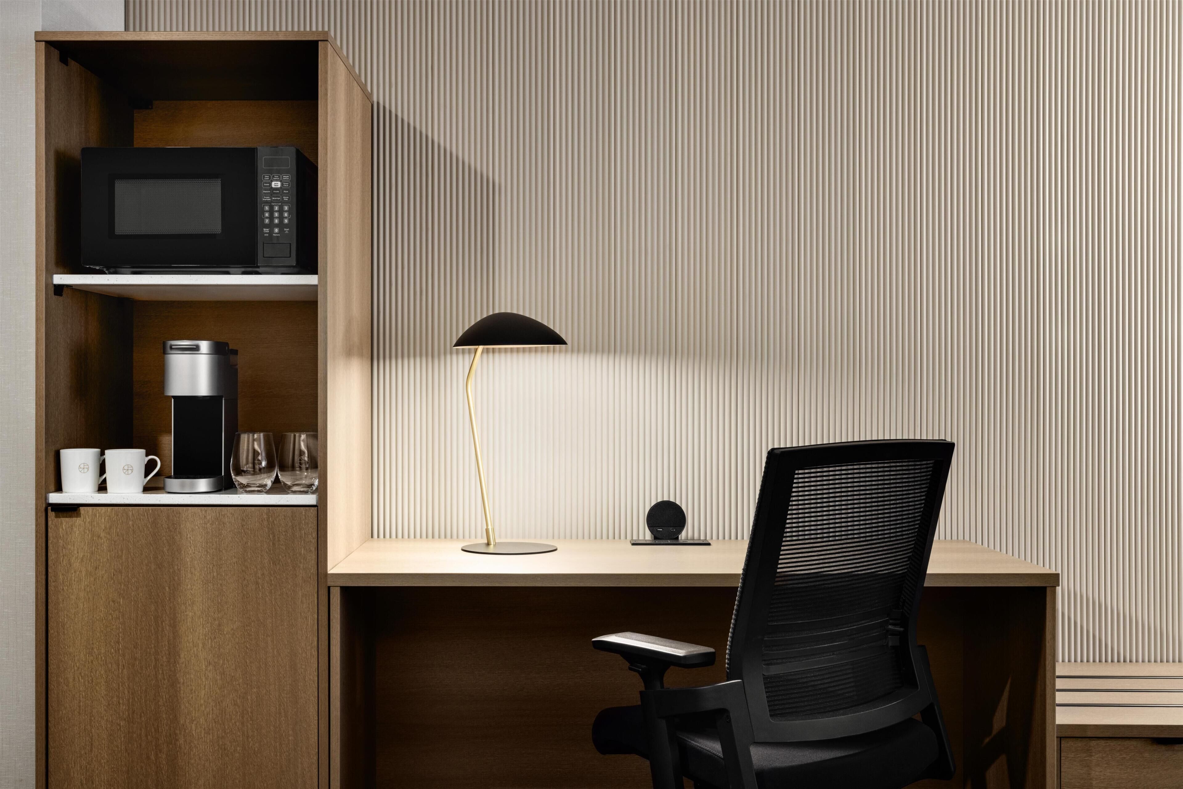 A modern office space with a wooden desk, a black office chair, a black lamp, a coffee maker, and a microwave on a shelf.