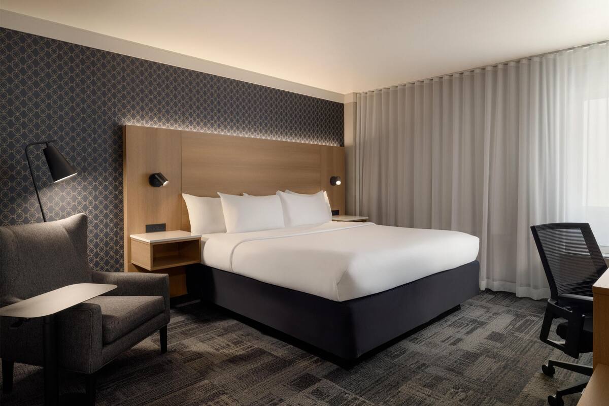 A hotel room with a large bed, a desk, a chair, a nightstand, and a lamp.