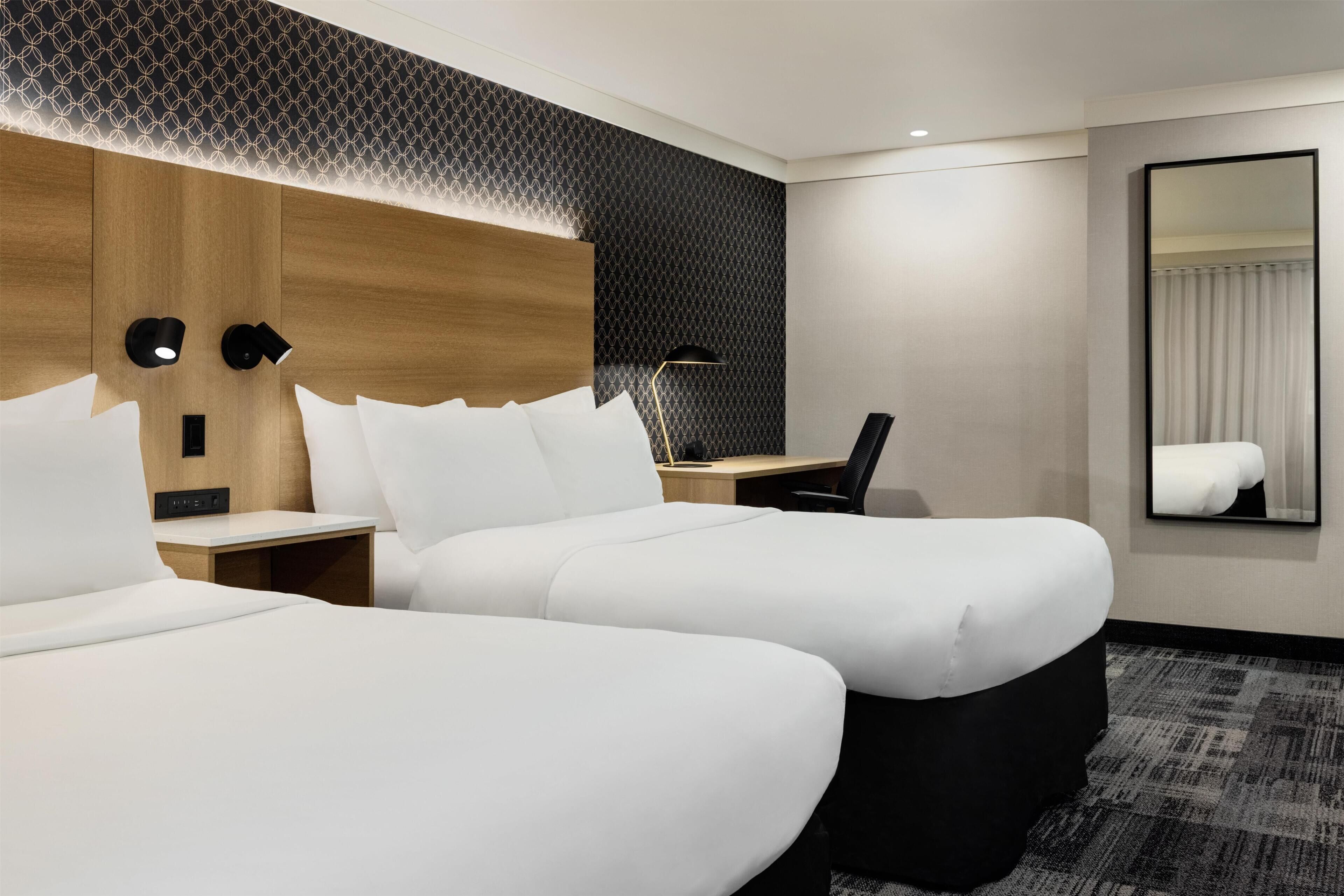 A hotel room with two beds, a desk, a chair, a mirror, and a patterned wall.