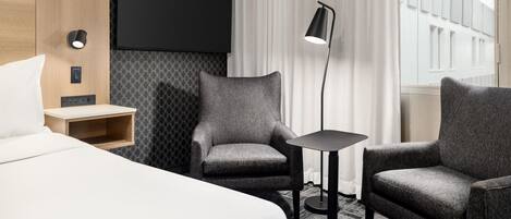 Habitación de hotel con cama, dos sillones, una mesita, televisor montado en la pared y una ventana con cortinas.