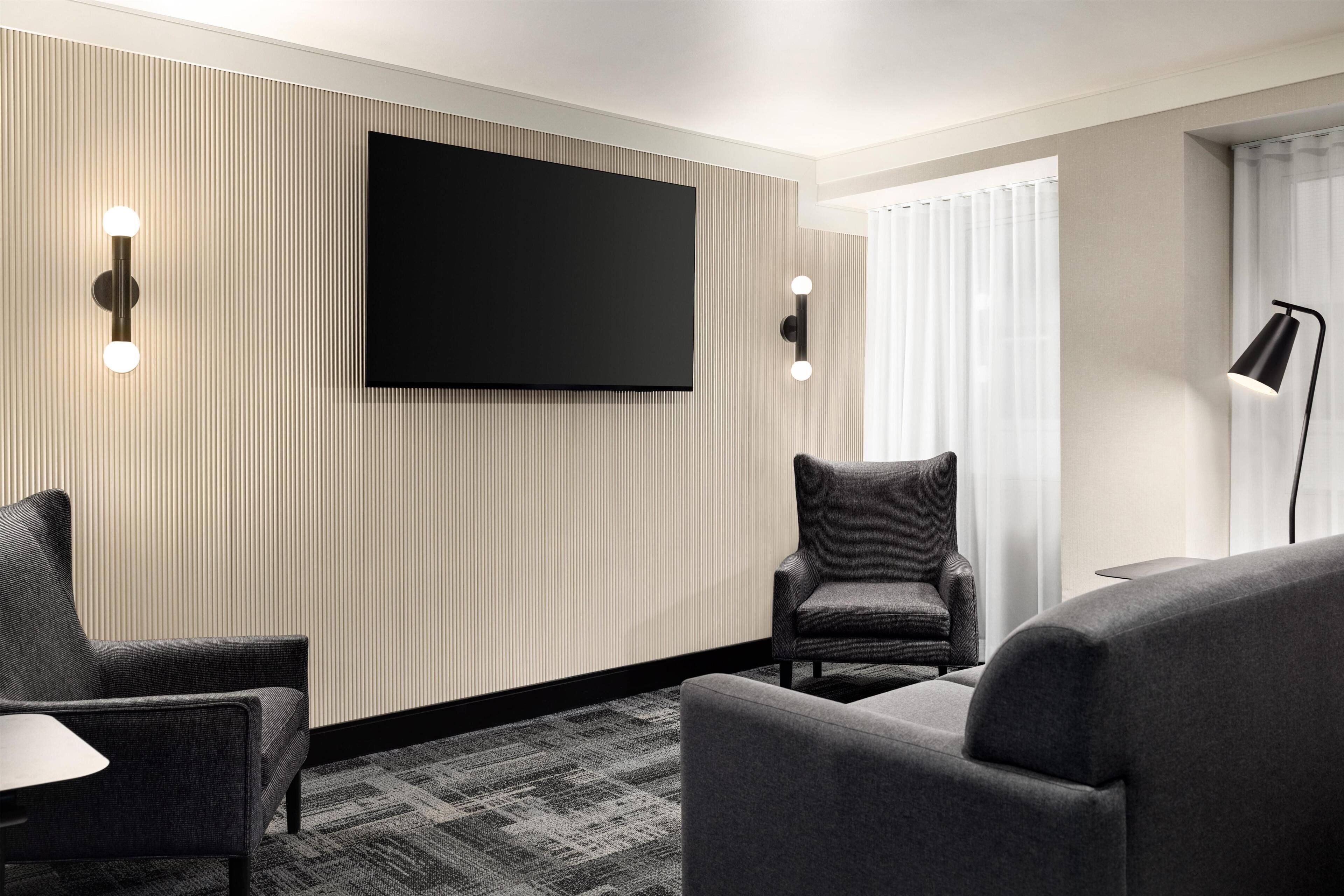 A modern hotel room with a flat-screen TV mounted on the wall, two armchairs, a small table, and a floor lamp.