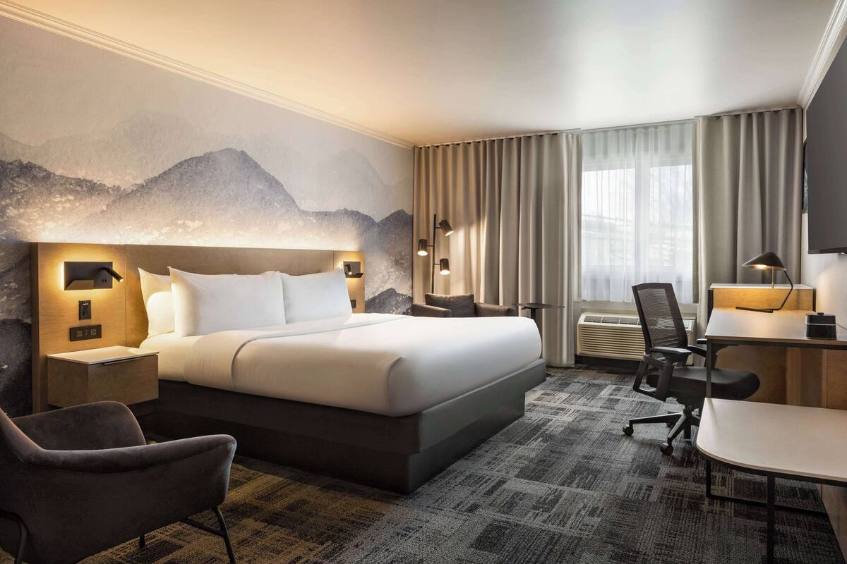 A hotel room with a large bed, a desk, a chair, a flat-screen TV, and a mountain mural on the wall.