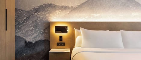 A hotel room with a bed, a nightstand, a wall-mounted light, and a mountain mural.