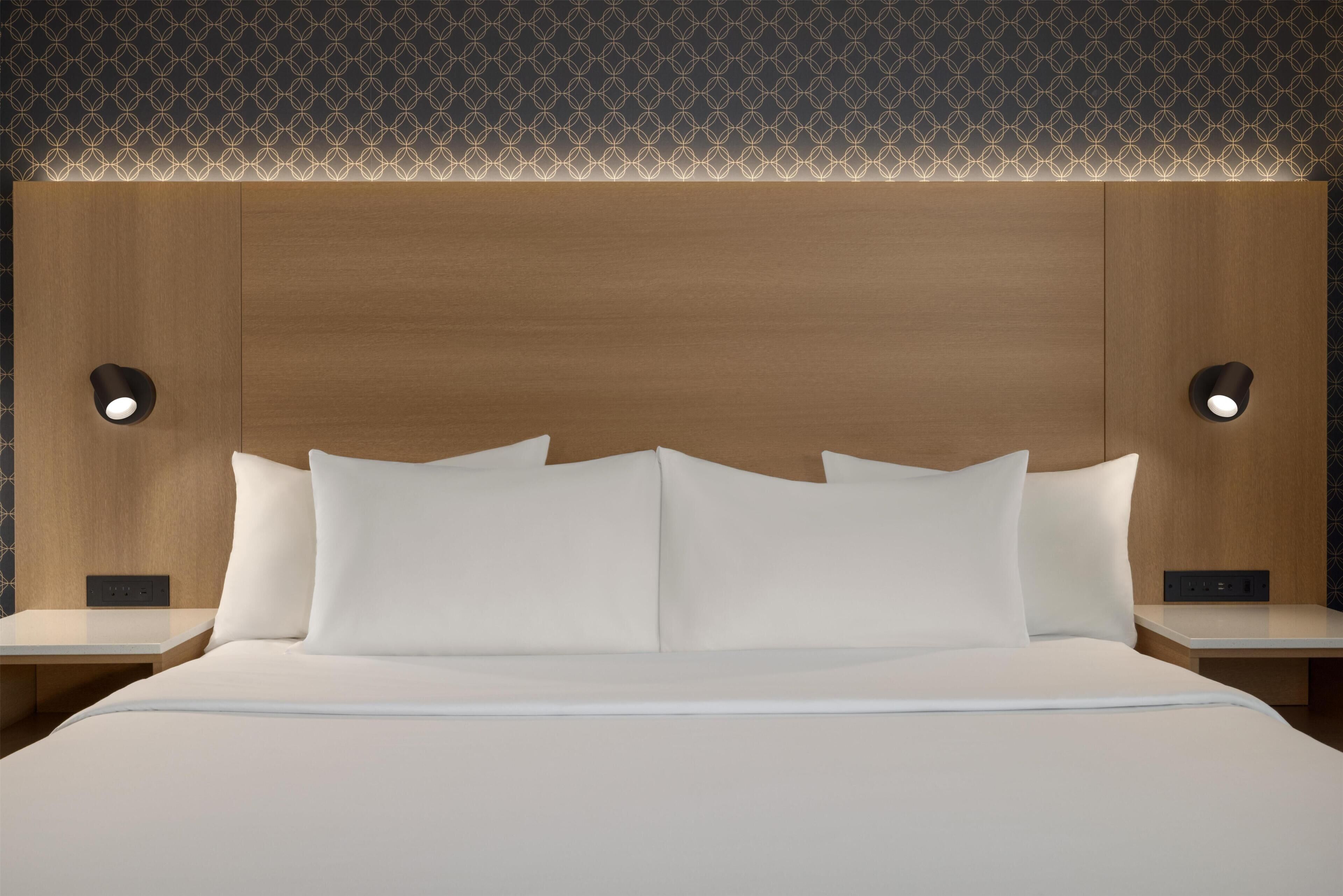 Habitación de hotel con cabecera de madera, dos mesitas de noche, ropa de cama blanca y lámparas de lectura fijadas a la pared.