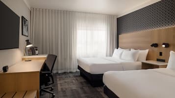 Une chambre d’hôtel avec deux lits, un bureau, une chaise, une lampe et une grande fenêtre avec des rideaux.
