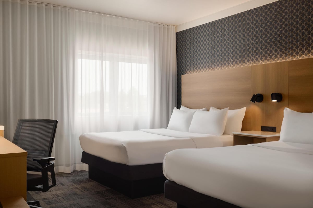 A hotel room with two beds, a desk, a chair, and a large window with sheer curtains.