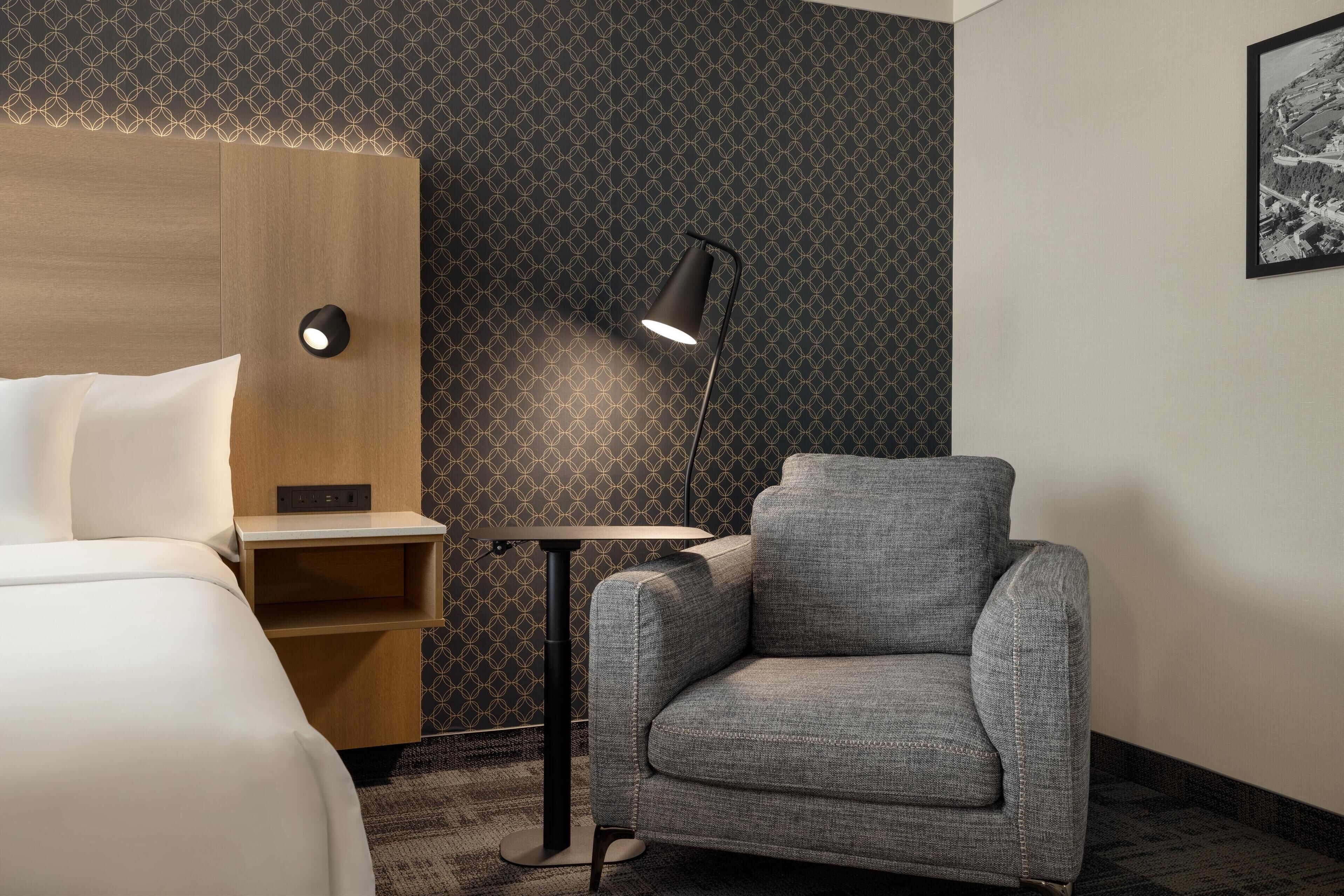 A hotel room with a bed, a grey armchair, a small table, a lamp, and a patterned wallpaper.