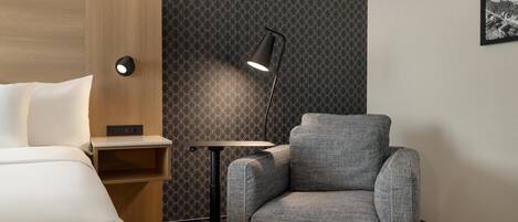A hotel room with a bed, a grey armchair, a small table, a lamp, and a patterned wallpaper.