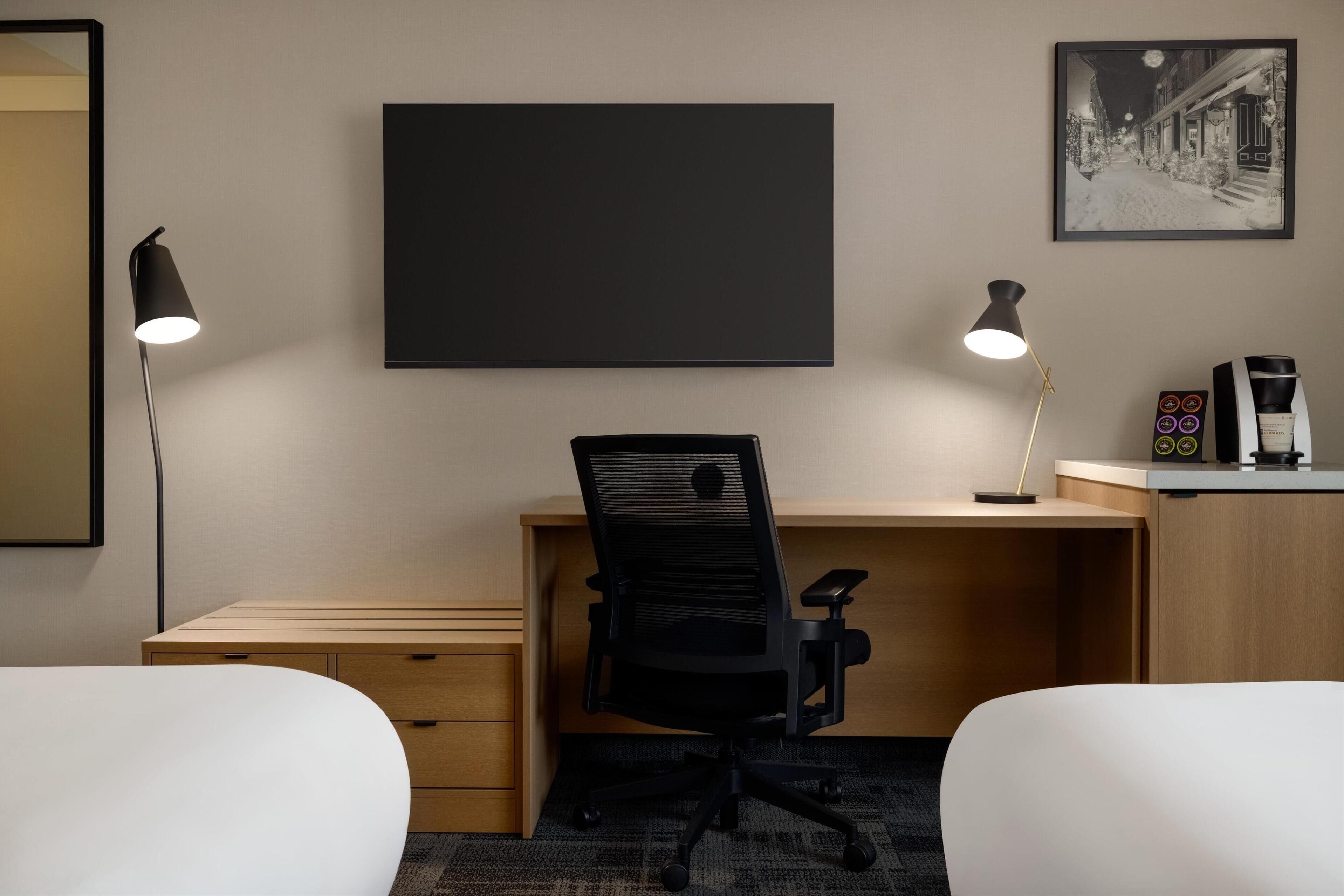 A hotel room with a wooden desk, two black office chairs, a large framed picture on the wall, and a coffee machine on the desk.