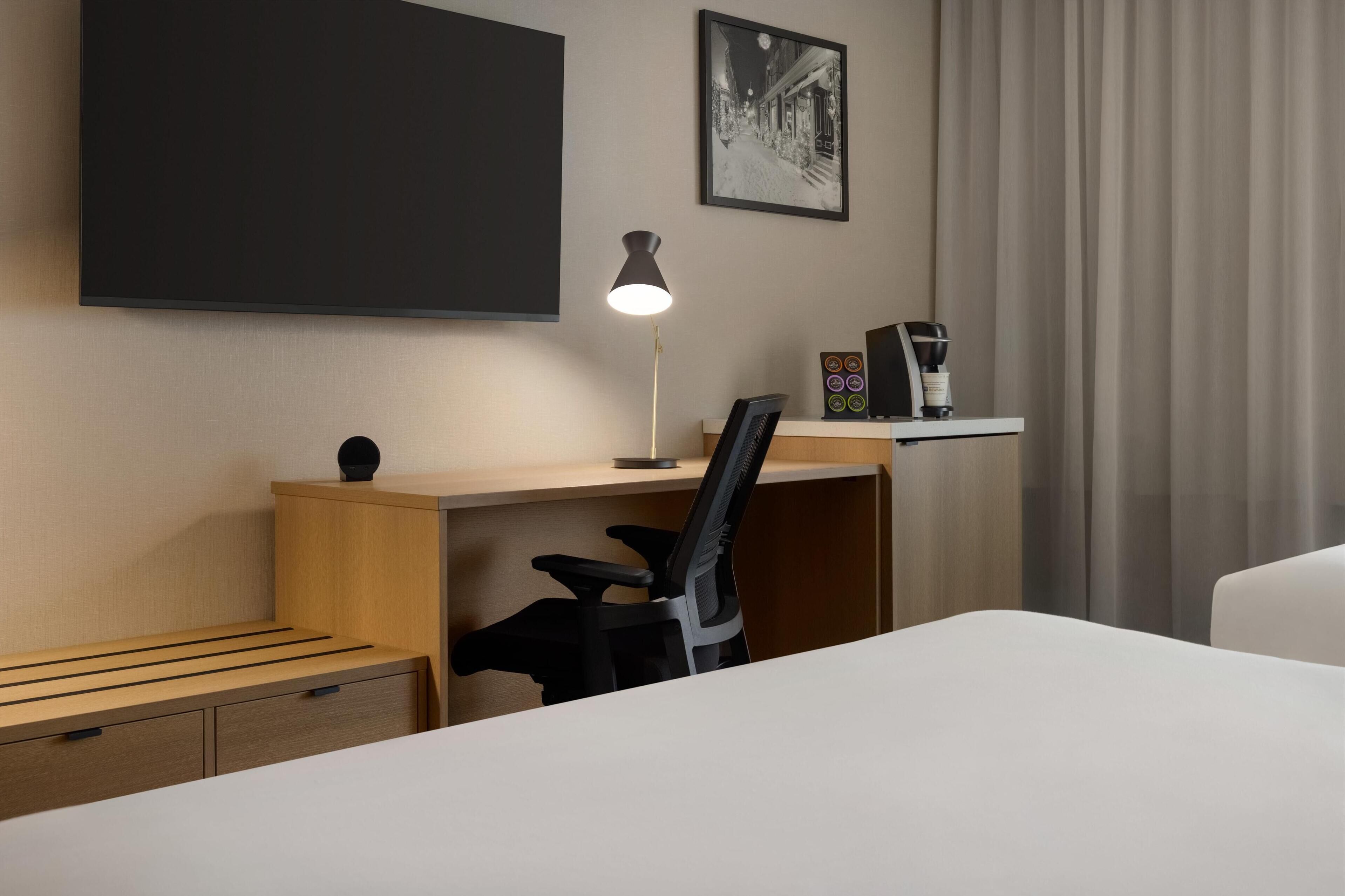 A hotel room with a bed, a desk with a chair, a lamp, a coffee machine, and a picture on the wall.
