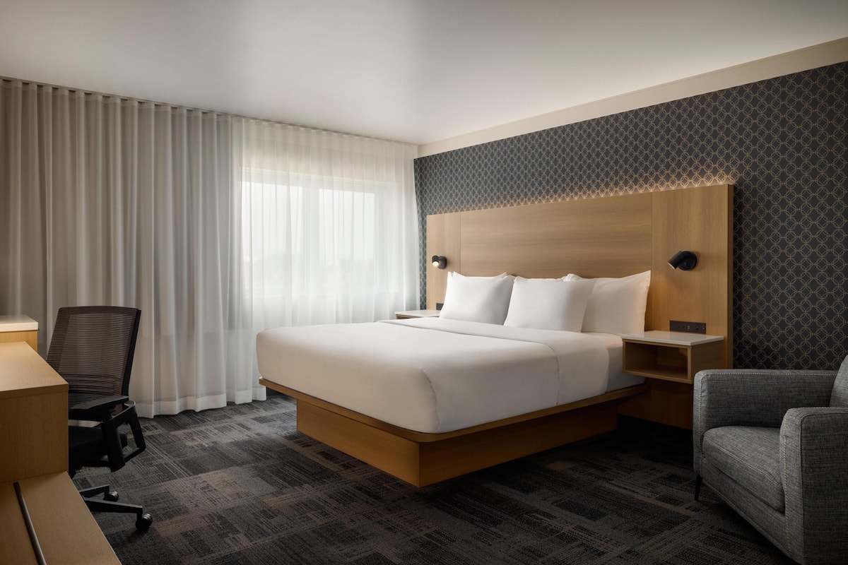 A hotel room with a large bed, a desk, a chair, and a window with curtains.