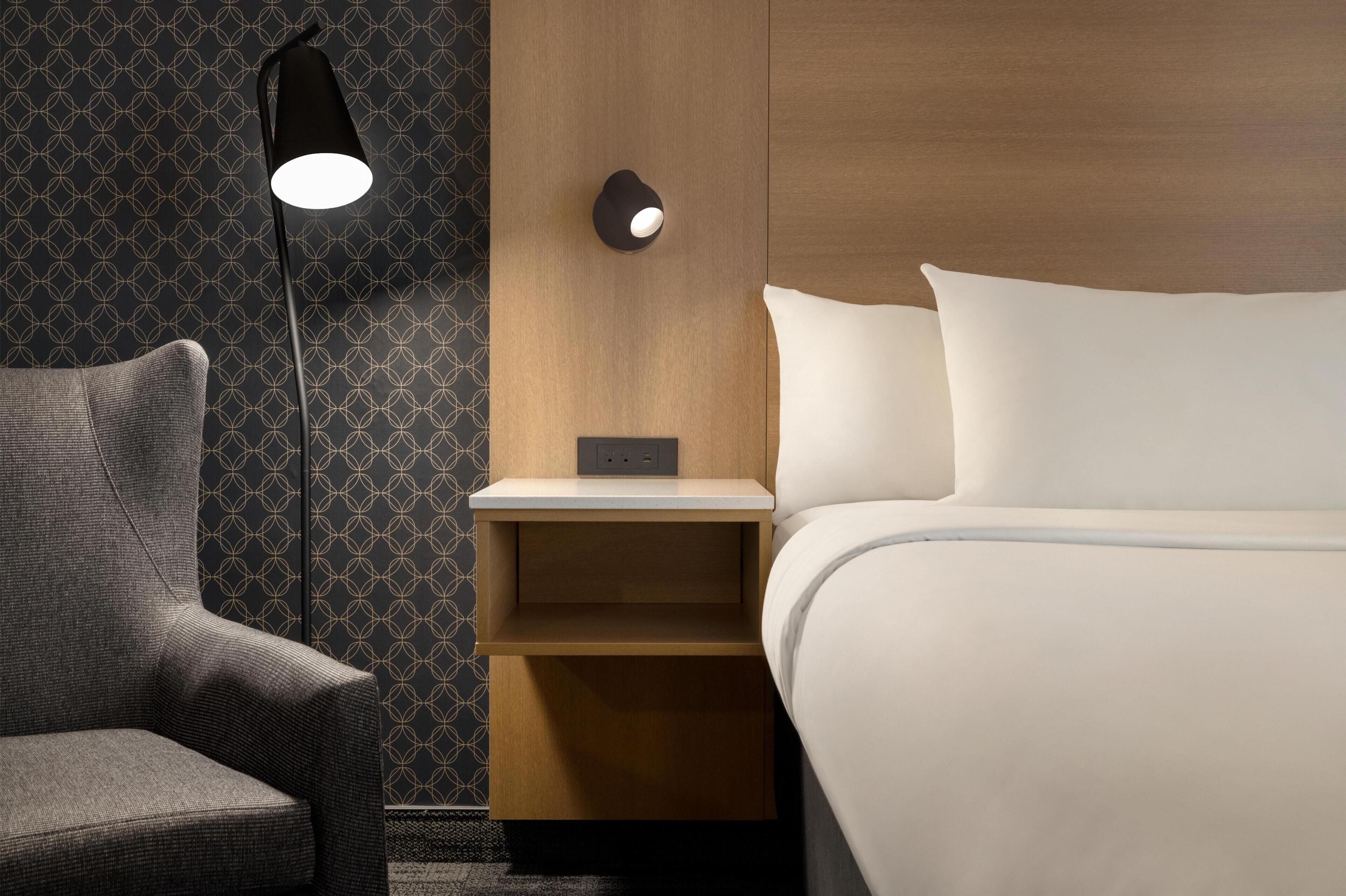 A hotel room with a bed, a nightstand, a lamp, and a chair.