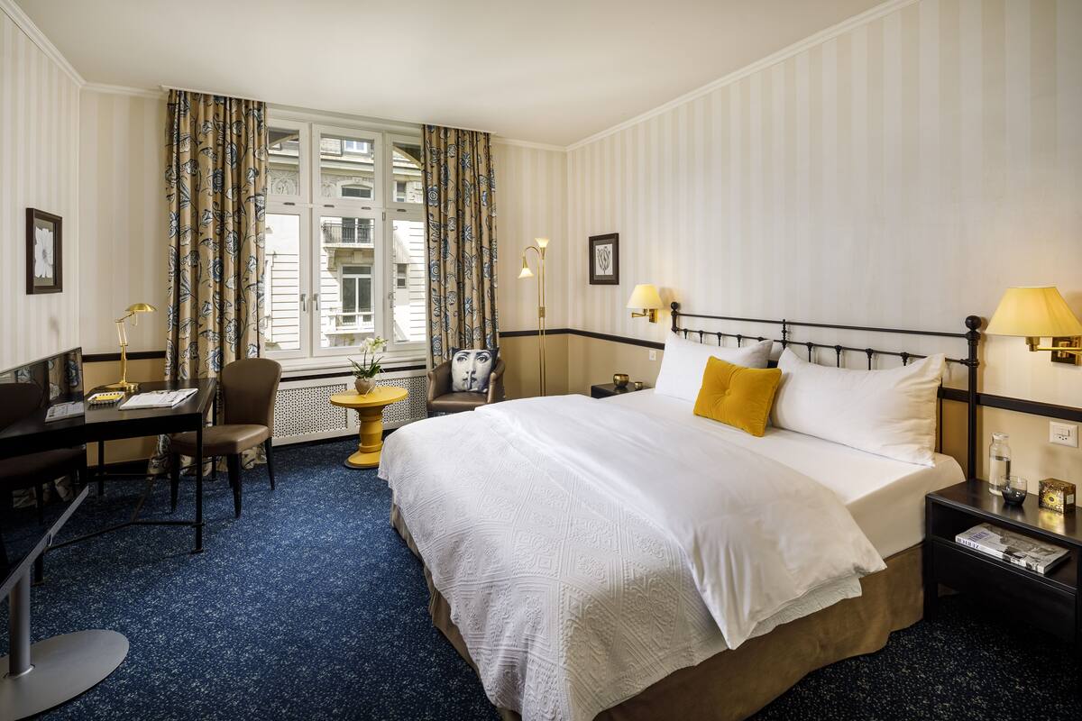A hotel room with a large bed, a desk, a chair, a small table, and a window with curtains.