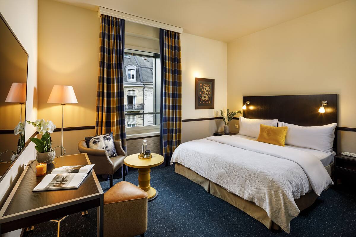 A hotel room with a bed, a desk, a chair, a small round table, and a window with curtains.