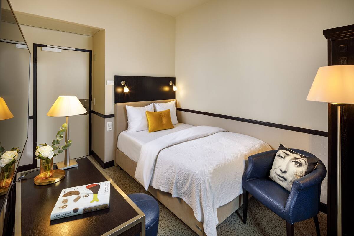 A hotel room with a bed, a chair, a desk with a lamp, and a mirror.