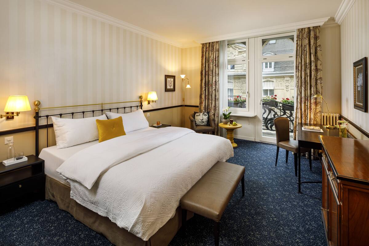 A hotel room with a large bed, bedside lamps, a bench, a desk, and a view of the outside balcony.