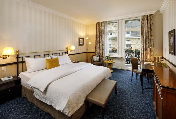 A hotel room with a large bed, bedside lamps, a bench, a desk, and a view of the outside balcony.