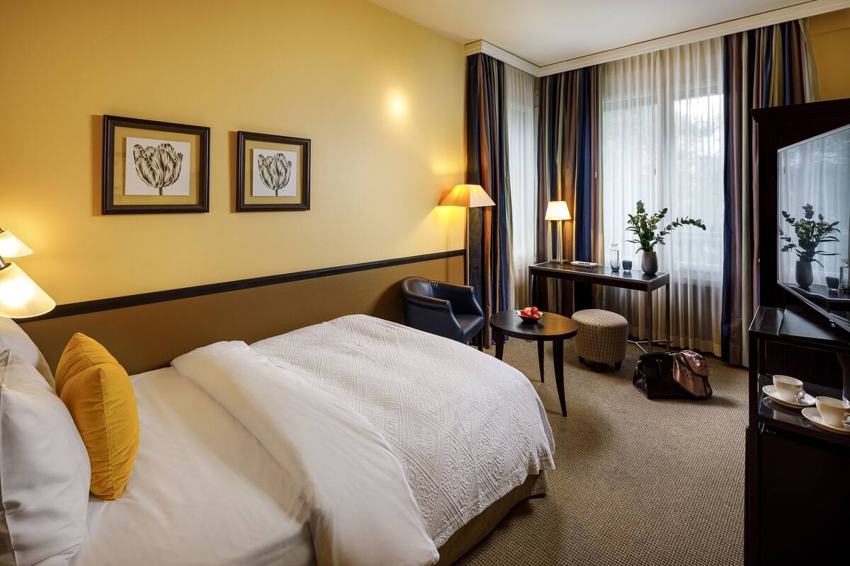 A hotel room with a large bed, two framed pictures on the wall, a desk with a lamp, a chair, a small table with a plant, and a window with curtains.