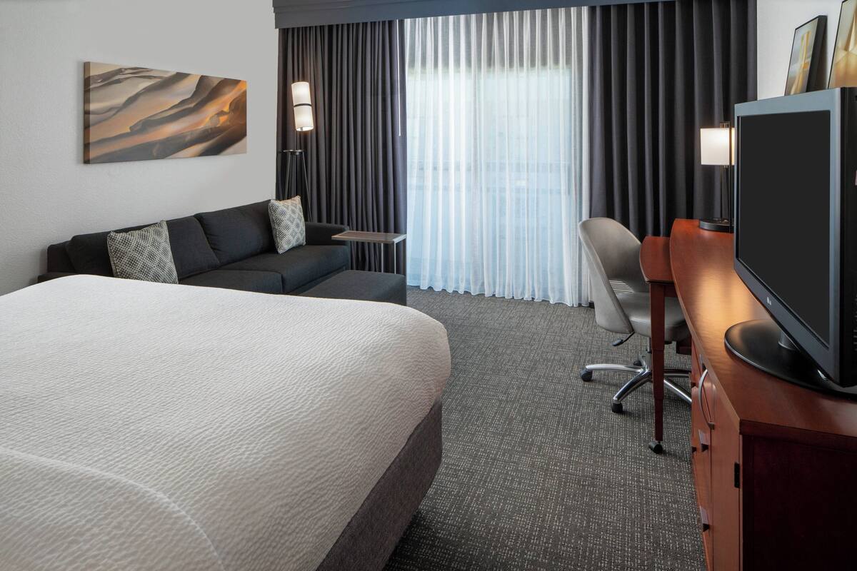 Ein Hotelzimmer mit einem großen Bett, einem Sofa, einem Schreibtisch mit Fernseher und einem Fenster mit Vorhängen.