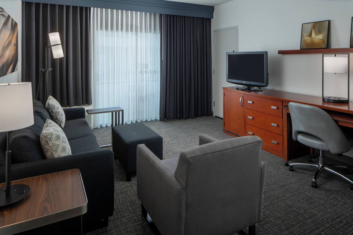 Ein Hotelzimmer mit Sofa, Hocker, Schreibtisch, Stuhl, Fernseher und Lampe.