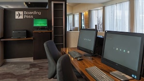 A modern workspace with two desks, each with a computer monitor, keyboard, and mouse. A 'Boarding Pass' sign is visible on the wall, and a television screen displays a landscape.