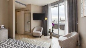 Habitación de hotel con una cama, dos sillones, un televisor, una mesita con una botella y una flor, y vistas a la ciudad a través de amplios ventanales.