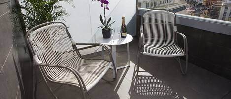 A balcony with two wicker chairs, a small table, a potted plant, and a bottle of champagne.