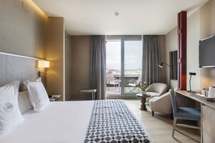 A hotel room with a large bed, a desk with a chair, a TV, and a balcony with a table and chairs.