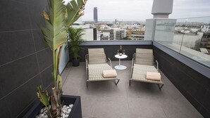 Una terraza en la azotea con dos sillones de mimbre, una mesita redonda con una botella y vasos, y una planta en maceta.