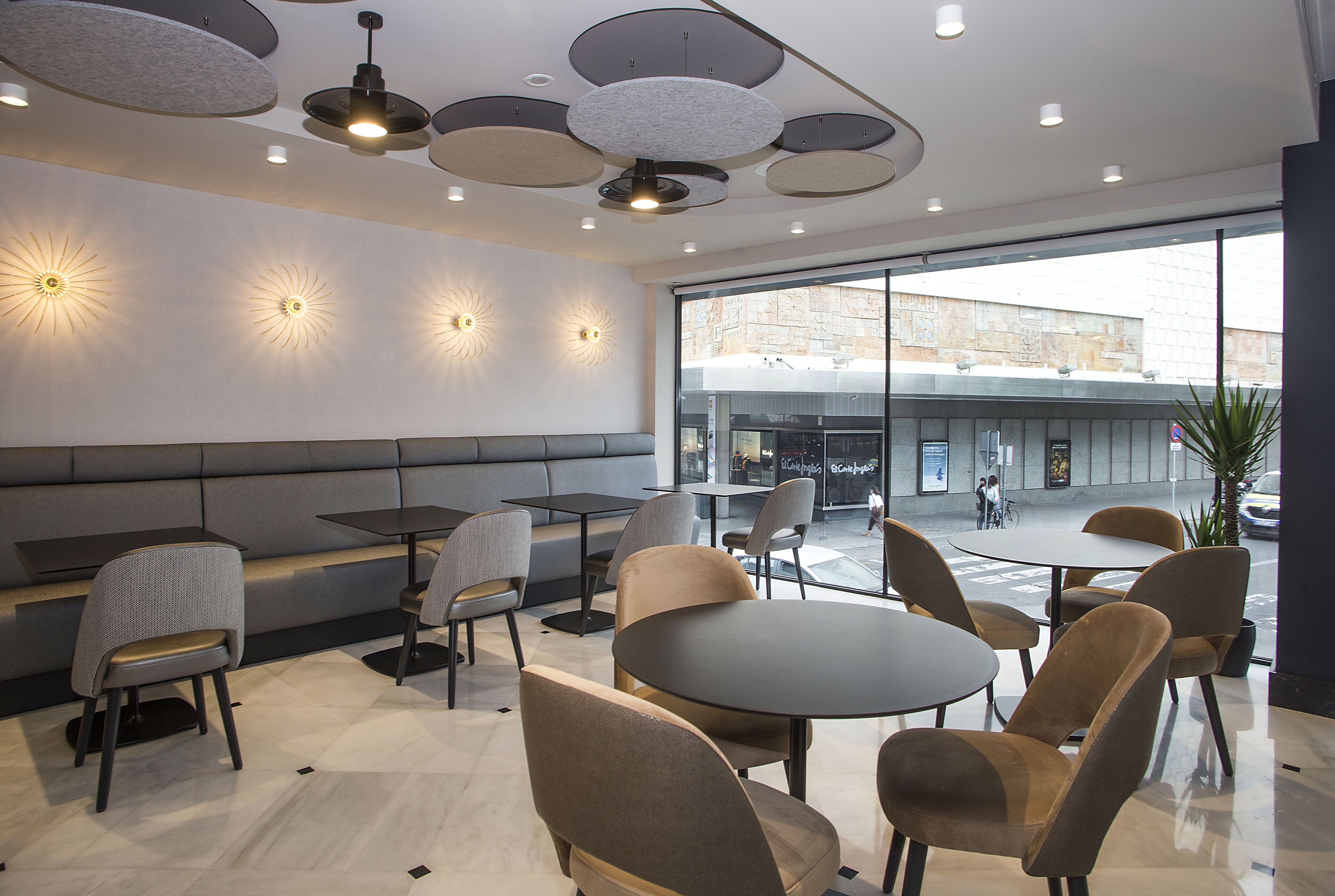 A modern cafe interior with seating arrangements, a large window offering an outdoor view, and contemporary lighting fixtures.