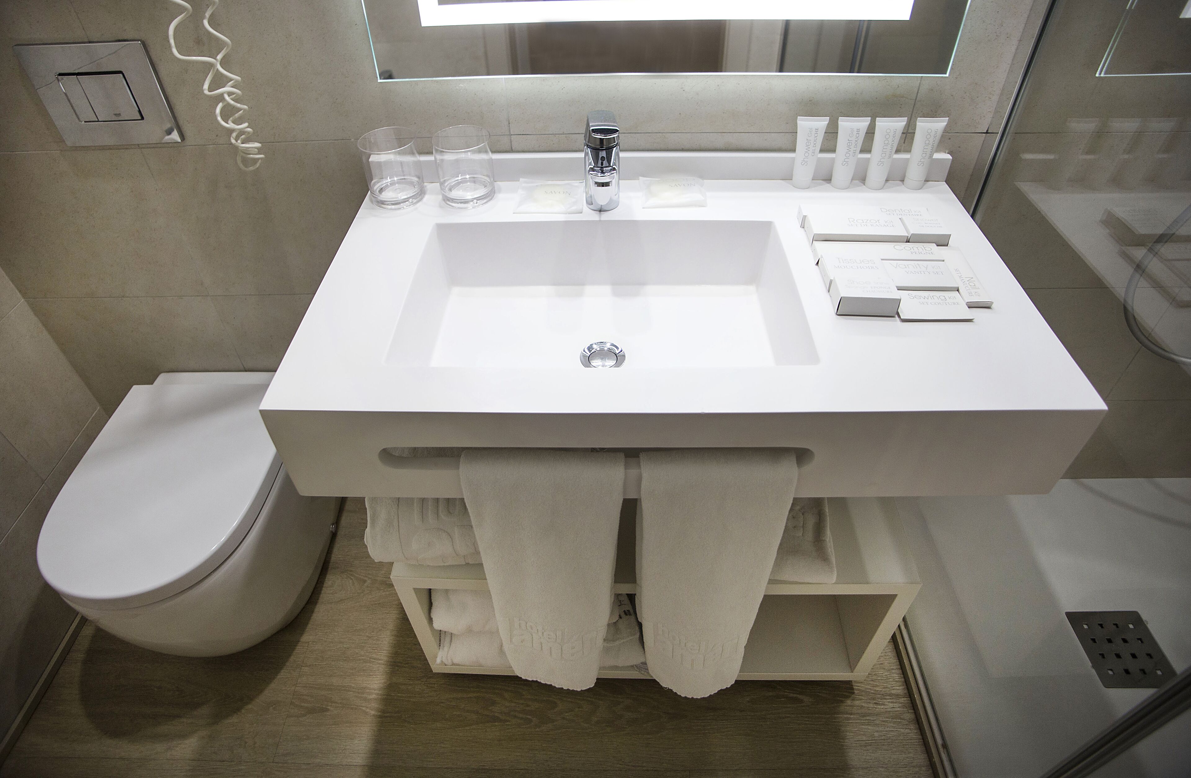 Un baño moderno con lavamanos, espejo y retrete blancos. Hay toallas colgadas en un perchero y un vaso de agua sobre el lavamanos.