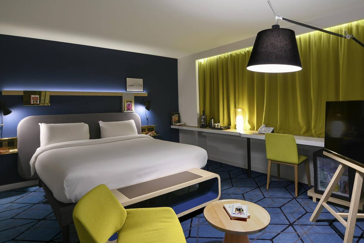 A hotel room with a bed, desk, chair, TV, lamp, and a small table with a book.