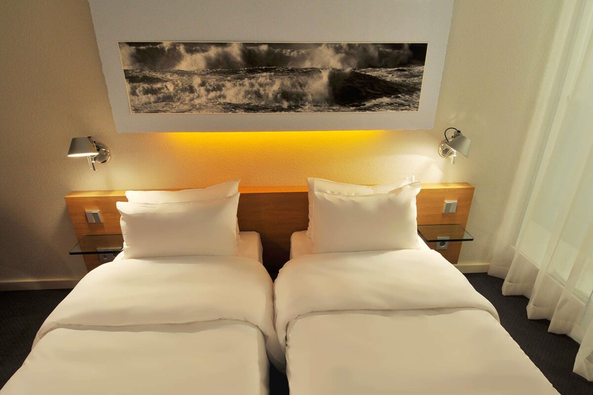 Een hotelkamer met twee bedden, een nachtkastje, een lamp en een ingelijst schilderij boven de bedden.
