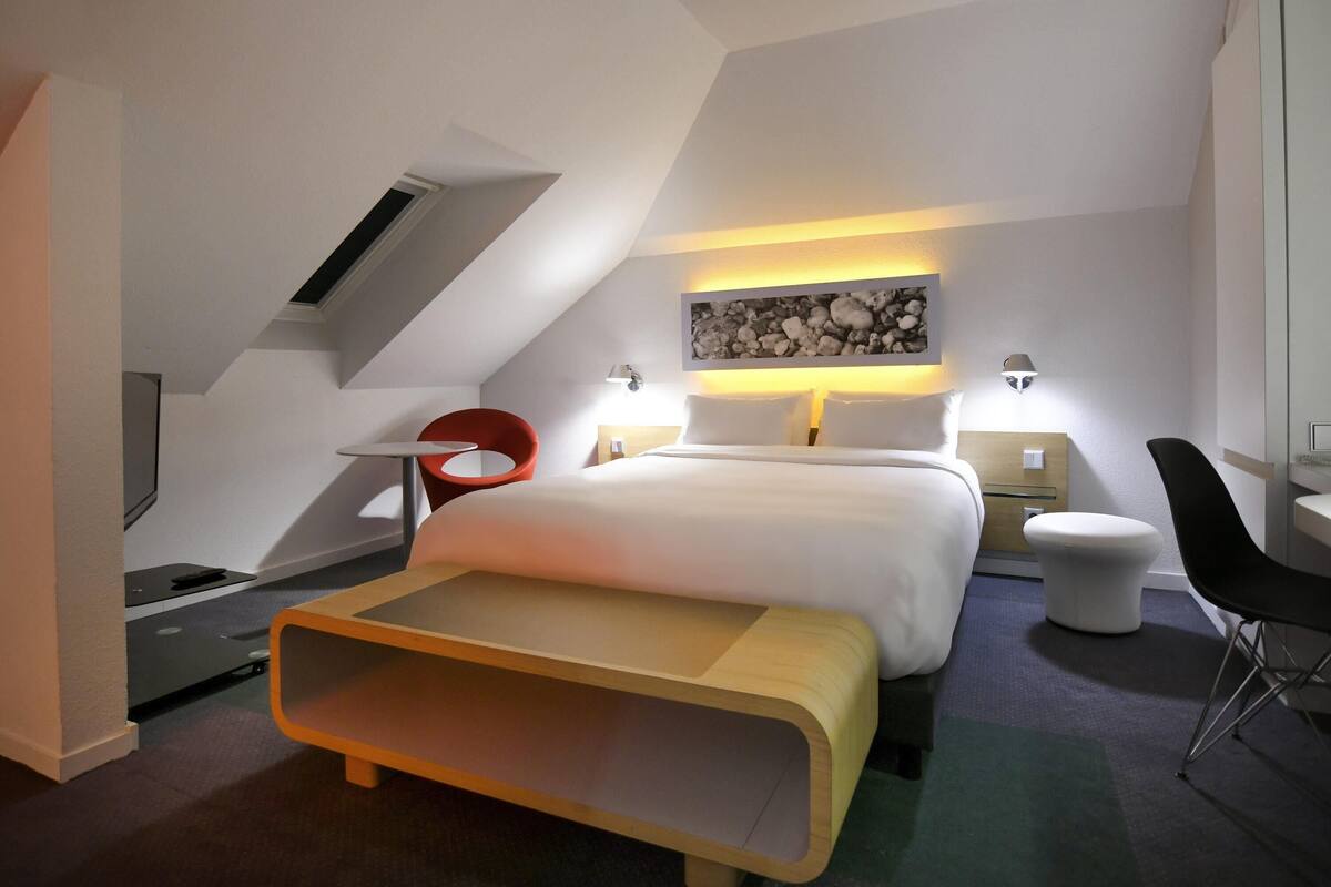 A modern hotel room with a bed, a desk, a chair, a small table, and a wall-mounted artwork.