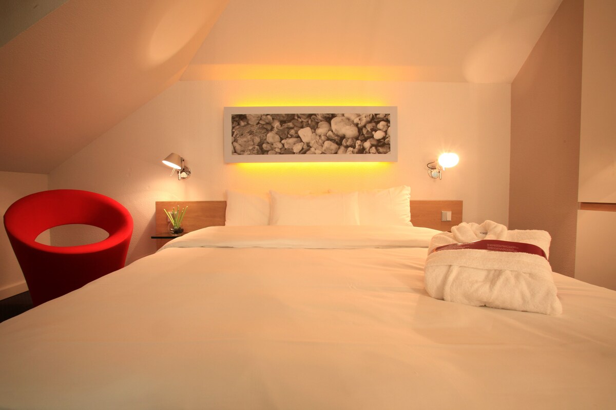 Une chambre d’hôtel avec un lit, un fauteuil rouge, une œuvre d’art accrochée au mur et deux appliques murales.