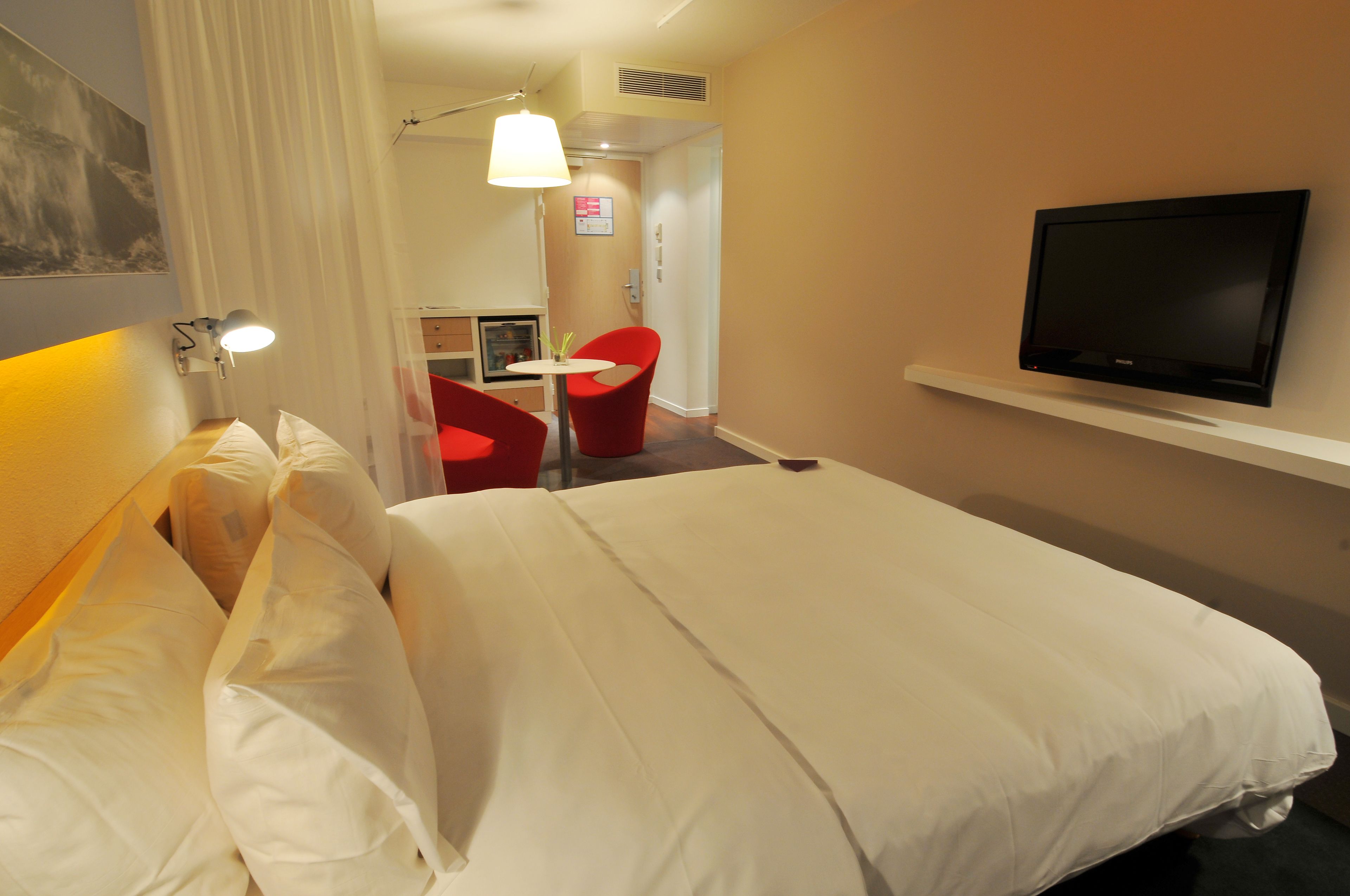 Une chambre d’hôtel avec un grand lit, un téléviseur fixé au mur, un bureau avec une chaise et, au fond, une petite kitchenette.