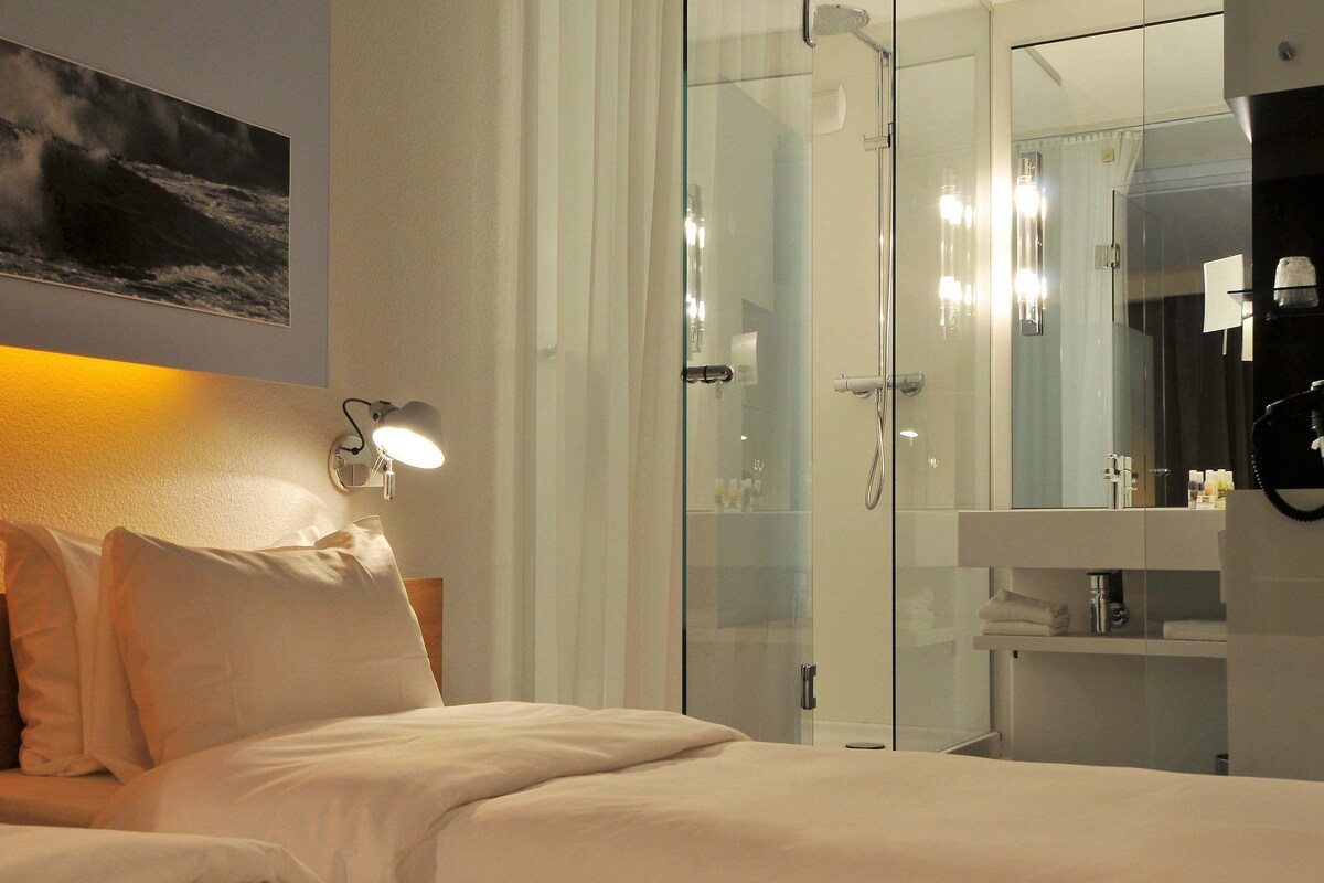 Une chambre d’hôtel comprenant un lit, une douche en verre, une salle de bain avec un miroir et une lampe fixée au mur.