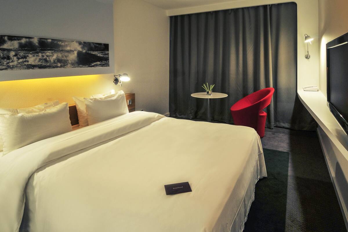 A hotel room with a large bed, a small table with a vase, a red chair, and a TV on the wall.