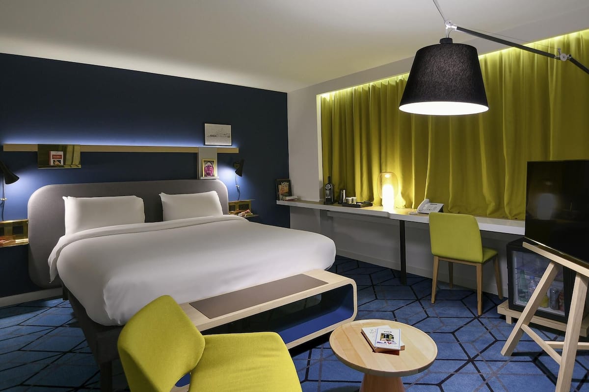 Una camera d'albergo con un letto, una scrivania, una sedia, una TV e un piccolo tavolino con un libro.