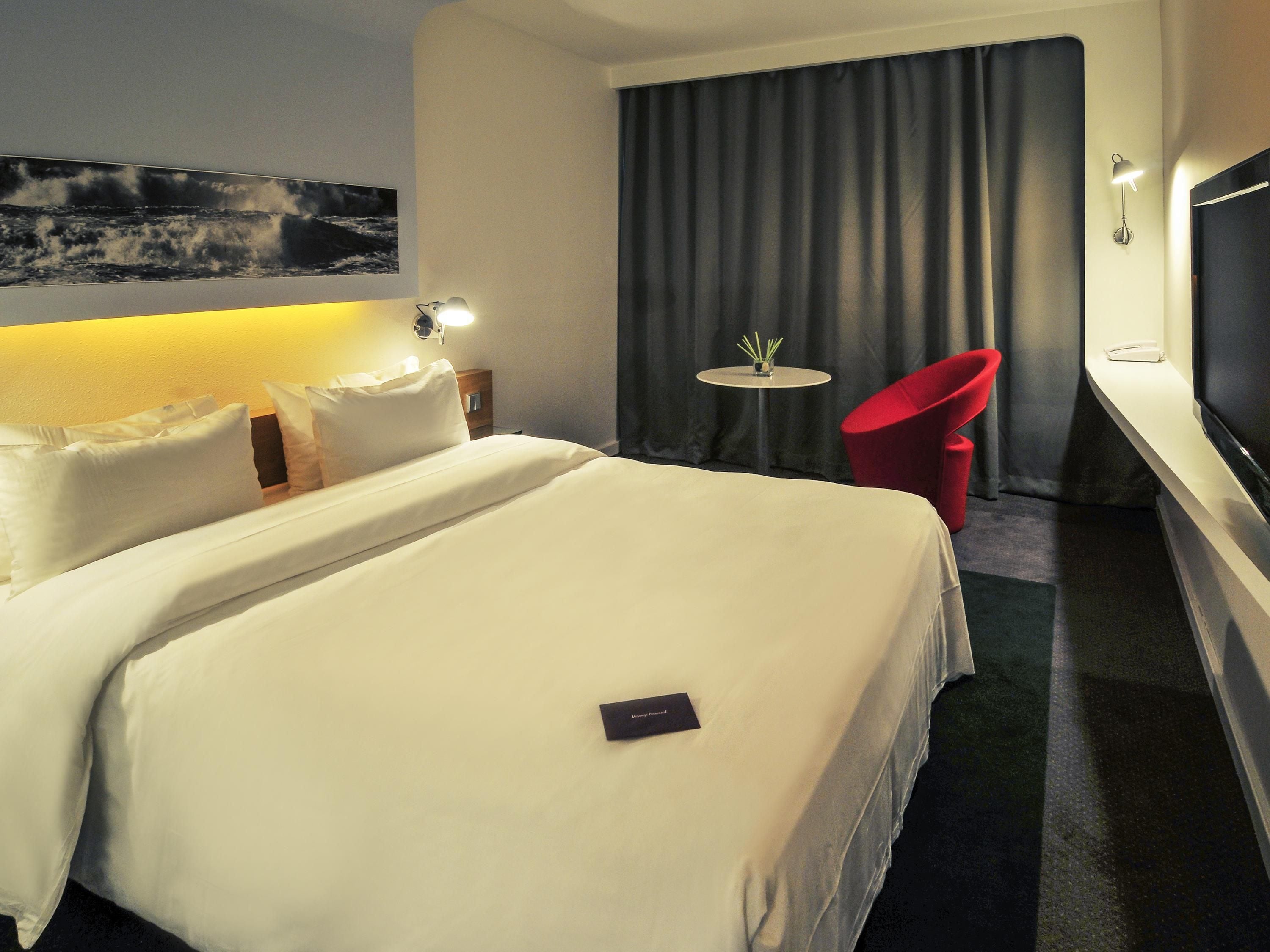 A hotel room with a large bed, a small table with a vase, a red chair, and a TV on the wall.